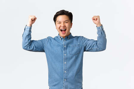 Excited happy asian man with braces feeling success over winning prize, fist pump and shouting yes, rejoicing, triumphing as becoming champion, standing white background celebratingの写真素材