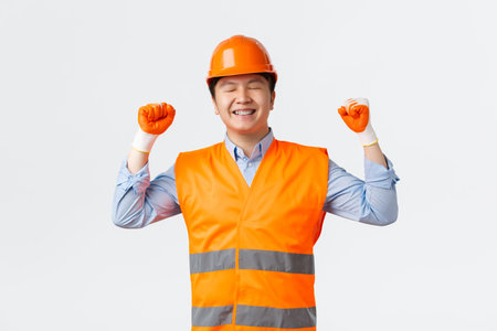 Building sector and industrial workers concept. Successful winning asian chief engineer finish work, looking delighted, fist pump in celebration, smiling pleased, wearing helmet and safety glovesの写真素材