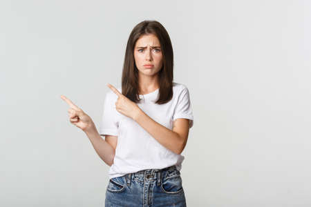 Nervous timid cute girl pointing fingers upper left corner and looking sad, having regretの写真素材
