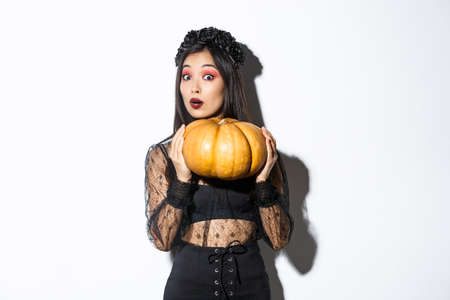 Portrait of woman lifting heavy pumpkin, getting ready for halloween, wearing witch costume, standing over white backgroundの写真素材