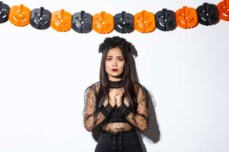 Image of scared and worried asian woman in witch costume looking concerned, wearing witch costume and standing against pumpkin bannersの写真素材