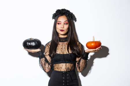 Thoughtful stylish woman in witch costume, picking pumpkin for halloween, making decision while...