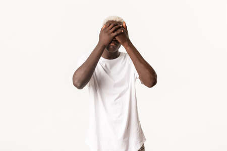 Portrait of gloomy and sad african-american blond guy, shut eyes with hands and looking miserable, standing white backgroundの写真素材