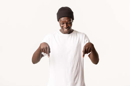 Portrait of happy and amused african-american handsome guy in beanie, looking and pointing fingers down at something interesting, white backgroundの写真素材
