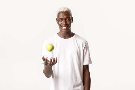Portrait of sassy handsome african-american blond guy, catching apple and smiling confident, standing white backgroundの写真素材