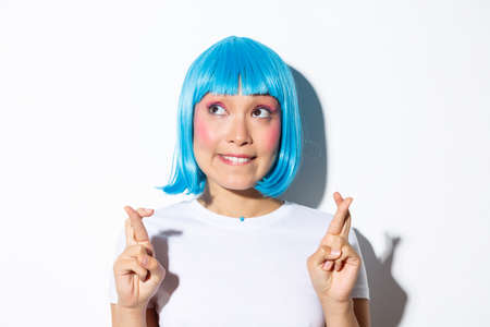 Cute asian girl making wish with crossed fingers, biting lip and looking tempted at upper left corner, wearing wig during holiday celebrationの写真素材