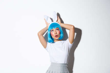 Portrait of coquettish asian girl in blue short wig and rabbit ears, looking up surprised, standing over white backgroundの写真素材