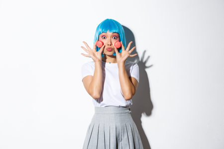 Portrait of silly asian girl in blue wig, glamour makeup, showing macaroons over face and pouting...