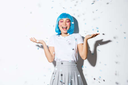 Portrait of happy asian girl in blue wig celebrating halloween, throwing confetti in the air, standing over white backgroundの写真素材