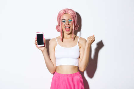 Image of beautiful party girl rejoicing, celebrating holiday, showing smartphone screen and shouting for joy, wearing pink wig and halloween outfitの写真素材