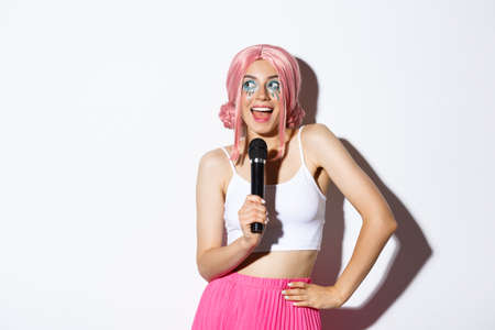 Image of beautiful smiling girl in pink wig, singing song in microphone, wearing halloween costume for party, standing over white backgroundの写真素材