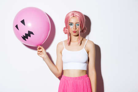 Image of sassy beautiful girl with pink wig and bright makeup, holding balloon at party, winking and looking coquettish, celebrating holidayの写真素材