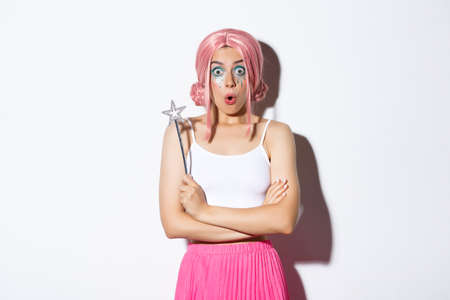 Portrait of surprised girl in pink wig, dressed as fairy for halloween party, holding magic wand and looking wondered, standing over white backgroundの写真素材