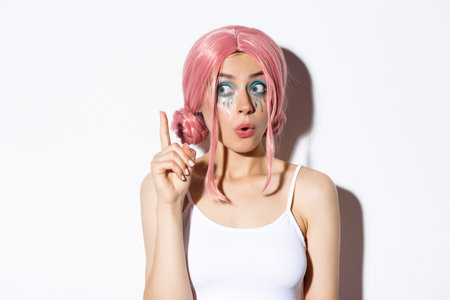 Close-up of woman in pink party wig and bright makeup, have idea, raising index finger in eureka...
