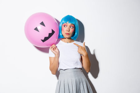 Beautiful asian girl celebrating halloween and wearing blue wig for party, pointing finger at balloon with scary faceの写真素材