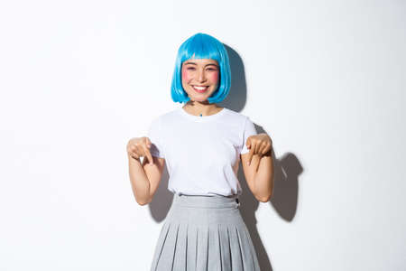 Cheerful asian woman in anime style outfit, wearing blue wig and smiling happy, pointing fingers down, showing advertisement, standing over white backgroundの写真素材