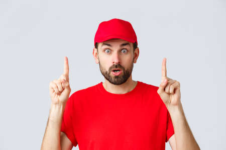 Online shopping, delivery during quarantine and takeaway concept. Surprised and impressed delivery guy, courier in red t-shirt and cap, pointing fingers up, gasping amazed, read interesting infoの写真素材