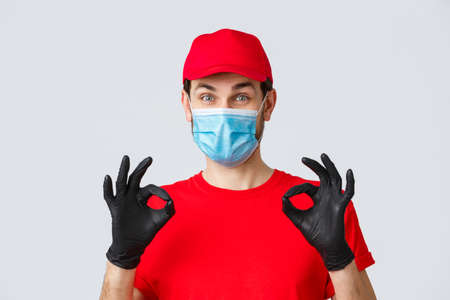 Contactless delivering, covid-19 and shopping concept. Cheerful, pleased courier in red uniform, cap and medical mask with gloves show okay, approval or guarantee gesture, recommend serviceの写真素材