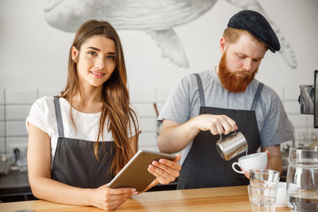 Coffee Business Concept - Cheerful baristas looking at their tablets for online orders in modern...