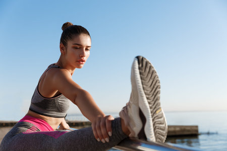 Close-up of attractive sportswoman holding leg on handrope, stretching on the seaside promenade....