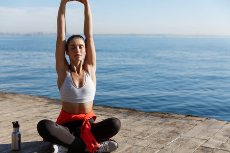 Outdoor shot of relaxed young woman meditating near the sea, practice yoga, sitting in asana on a...