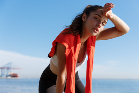 Close-up of attractive young fitness woman taking a break during jogging, panting and wiping sweat...