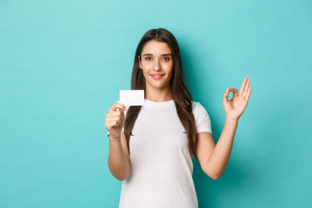 Portrait of satisfied brunette girl in white t-shirt, recommend bank, showing credit card and okay...