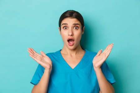 Coronavirus, social distancing and health concept. Close-up of alarmed and anxious female doctor, spread hands sideways and drop jaw, looking worried at camera, standing over blue backgroundの写真素材