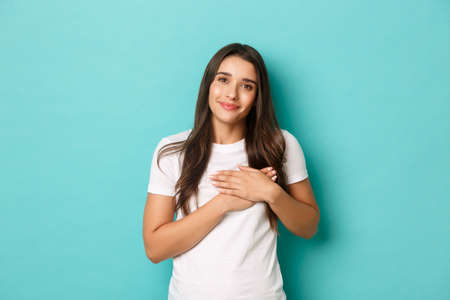 Portrait of attractive brunette girl in white t-shirt, thanking for something, holding hands on heart and looking with gratitude, standing over blue backgroundの写真素材