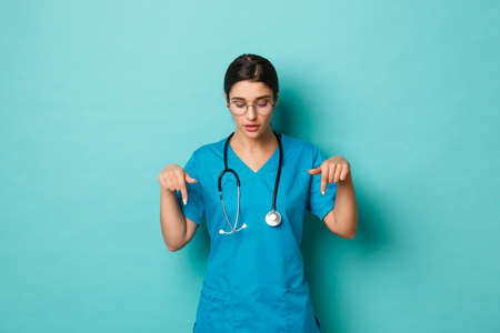 Coronavirus, pandemic and social distancing concept. Image of thoughtful female doctor in scrubs, looking and pointing fingers down curious, standing over blue backgroundの写真素材