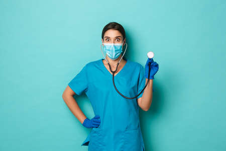 Covid-19, pandemic and medicine concept. Surprised female doctor in medical mask, gloves and scrubs, holding stethoscope and looking amazed, standing over blue backgroundの写真素材