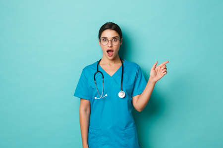 Coronavirus, pandemic and social distancing concept. Amazed female doctor in glasses and scrubs, pointing finger right at copy space, looking astonished, standing over blue backgroundの写真素材