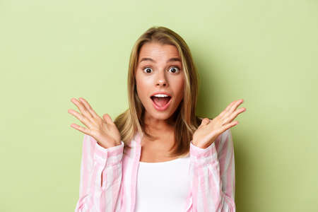 Close-up of attractive blond woman looking surprised, spread hands sideways and open mouth impressed, standing over green backgroundの写真素材