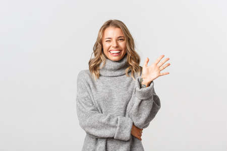 Image of friendly happy woman in grey sweater saying hello, smiling and waving hand to greet person, standing over white backgroundの写真素材