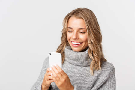Close-up of attractive modern girl with blond short hairstyle, wearing grey sweater, using mobile phone and smiling, standing over white backgroundの写真素材
