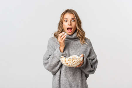 Image of amazed blond girl watching movie, looking excited, eating popcorn, standing over white backgroundの写真素材