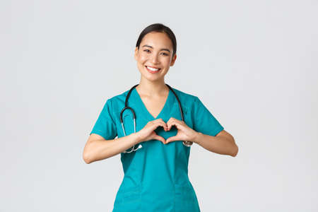 Covid-19, healthcare workers, pandemic concept. Lovely caring asian doctor, female nurse in scrubs showing heart gesture and smiling, taking care of patients with love, white backgroundの写真素材