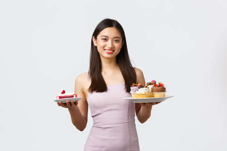 Lifestyle, holidays, celebration and food concept. Beautiful asian woman in dress holding plates with desserts, suggesting cake or plate with cupcakes, standing white backgroundの写真素材