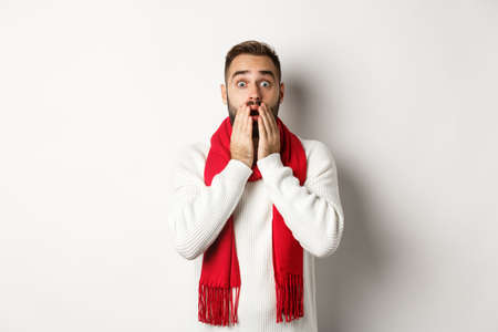 Christmas holidays and celebration concept. Handsome bearded guy in red scarf and sweater looking shocked, impressed with advertisement, white backgroundの写真素材