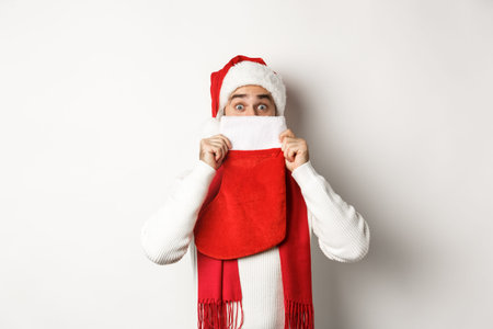 Winter holidays and shopping concept. Cheerful caucasian guy in Santa hat hiding face behind...