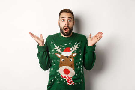 New Year party and winter holidays concept. Surprised bearded guy gasping amazed, standing in Christmas sweater, white backgroundの写真素材
