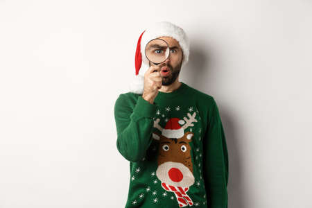 Christmas and holidays concept. Curious guy looking at something through magnifying glass, standing in Santa hat with xmas sweater, standing over white backgroundの写真素材