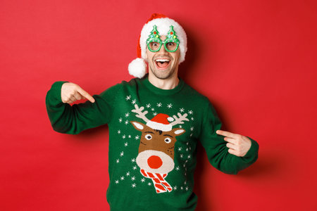 Image of happy man in party glasses and santa hat, pointing at his christmas sweater and smiling, standing over red backgroundの写真素材