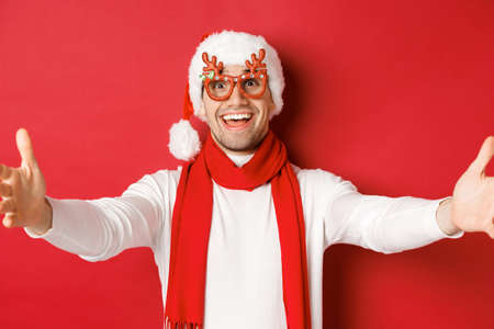 Concept of christmas, winter holidays and celebration. Close-up of happy smiling man in party glasses and santa hat, spread hands sideways for hug, greeting guests, red backgroundの写真素材