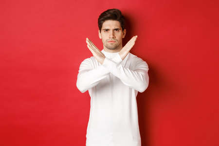 Image of serious and angry man in white sweater, express storng disapproval, showing cross sign to stop something bad, forbid action, standing over red backgroundの写真素材