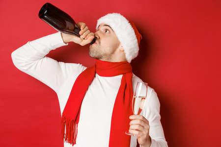 Concept of winter holidays, christmas and lifestyle. Man getting drunk on new year party, drinking champagne from bottle, wearing santa hat, standing over red backgroundの写真素材