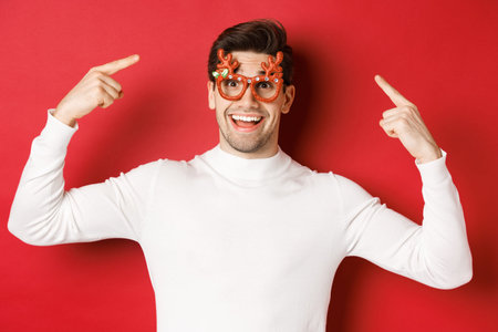 Handsome happy guy in white sweater, pointing at his christmas party glasses, celebrating new year...