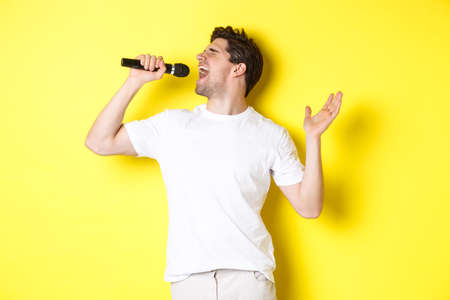 Young man singer holding microphone, reaching high note and singing karaoke, standing over yellow backgroundの写真素材
