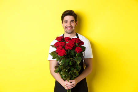 Smiling florist in black apron holding flowers, selling bouquet of roses, standing over yellow backgroundの写真素材