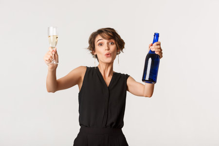 Cheerful elegant woman having fun on party, celebrating something, dancing with bottle and glass of champagne, standing over white backgroundの写真素材